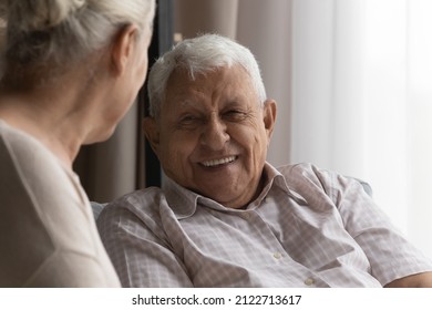 Happy elderly 80s man talking to wife, female carer at home, resting on couch, speaking to woman with toothy smile. Later life, old age, elderly care, marriage concept