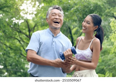 Happy elderly Asian man lifting up dumbbell in green park. Joyful senior enjoys exercising to get strength with personal trainer at outside garden. Sport woman giving advice on healthy exercise. - Powered by Shutterstock