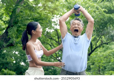 Happy elderly Asian man lifting up dumbbell in green park. Joyful senior enjoys exercising to get strength with personal trainer at outside garden. Sport woman giving advice on healthy exercise. - Powered by Shutterstock