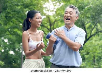 Happy elderly Asian man lifting up dumbbell in green park. Joyful senior enjoys exercising to get strength with personal trainer at outside garden. Sport woman giving advice on healthy exercise. - Powered by Shutterstock