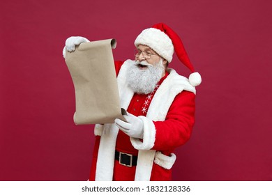 Happy elderly bearded Santa Claus wearing costume, hat, glasses, holding parchment roll reading letter wish list preparing for xmas eve isolated on red background. Merry Christmas wishlist ad concept. - Powered by Shutterstock