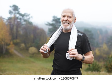 Happy Elderly Caucasian Male With Stubble Choosing Active Lifstyle, Smiling Broadly After Intensive Morning Cardio Workout In Park, Wiping Sweat Using White Towel. Health, Well Being And Energy