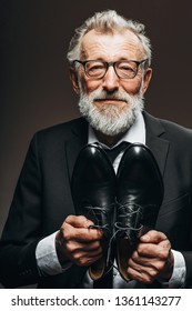 Happy Elderly Cobbler In Elegant Suit Looking At Camera While Demonstrating Bespoke Leather Shoes Near His Chest, Offering To Appreciate His Work