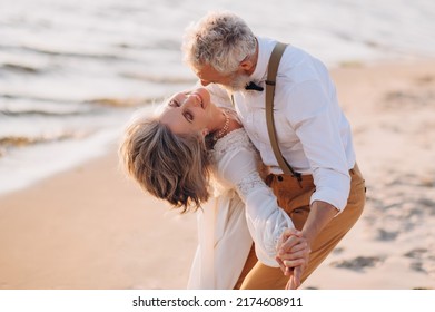 Happy Elderly Couple Dancing On The Beach On A Sunny Day. The Groom Dances With The Bride On The Wedding Day.