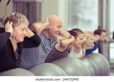 Happy Elderly Couple Exercising In A Pilates Class At The Gym With Three Other Younger People Toning And Strengthening Their Muscles Using Gym Balls, Focus To The Senior Man And Woman