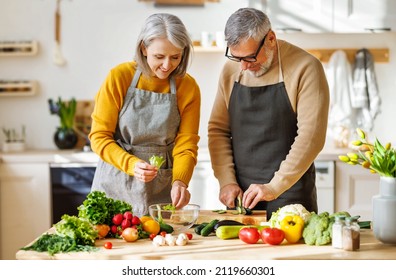 Happy elderly couple smiling husband and wife in aprons prepare salad together at kitchen table, chopping variety of colorful vegetables, trying to maintain healthy lifestyle eating vegetarian food