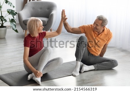 Happy elderly couple in sportswear sitting on fitness mat in living room, giving each other high five and smiling, training together at home, celebrating success, sports lifestyle for seniors