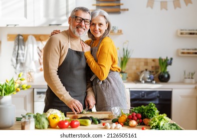 Happy Elderly Family Couple Wife And Husband Embracing And Smiling At Camera While Preparing Vegetarian Dish From Colorful Variety Of Vegetables And Herbs Lying On Kitchen Table In Front Of Them