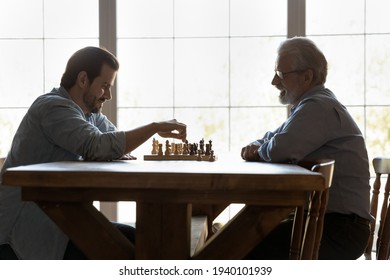 Happy Elderly Father Sit At Table Play Chess With Adult Grownup Son. Smiling Young Caucasian 30s Man Have Fun Enjoy Logic Board Game Set Or Match With Older 60s Dad On Family Weekend At Home.