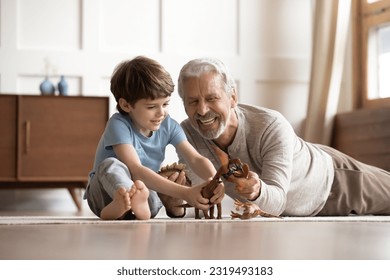 Happy elderly grandfather lying on floor at home have fun play toys with small 6s grandson together. Smiling mature grandparent feel playful engaged in funny activity with little boy child on weekend.
