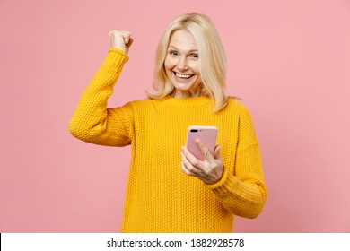 Happy Elderly Gray-haired Blonde Woman Lady 40s 50s Years Old In Yellow Sweater Using Mobile Cell Phone Typing Sms Message Doing Winner Gesture Isolated On Pastel Pink Background Studio Portrait