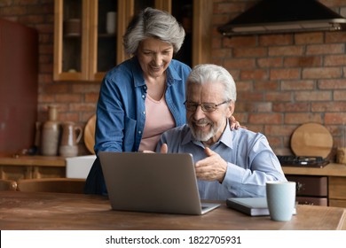 Happy Elderly Husband And Wife Sit At Table At Home Kitchen Look At Laptop Screen Reading Good News Online. Smiling Mature 70s Man And Woman Excited About Pleasant Bank Message Or Notice On Internet.