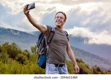 A Happy Elderly Male Tourist 45-50 Years Old With A Backpack Takes A Selfie Against The Backdrop Of A Mountain Landscape.