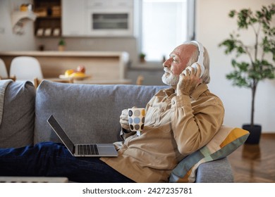 Happy elderly man at home using laptop. Happy elderly man relaxing on a sofa at home and listening to music on laptop with headphones, while drinking coffee. - Powered by Shutterstock