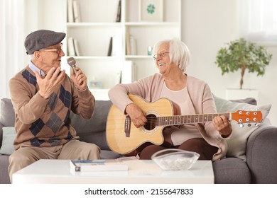 Happy elderly man with a microphone at home and an elderly woman playing an acoustic guitar seated on a sofa - Powered by Shutterstock