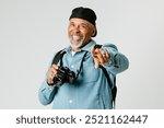 Happy elderly man photographer with a camera standing against white background. Cheerful elderly man pointing at camera. Senior man photographer traveling and taking photos of joyful moment.