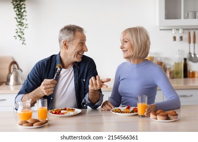 Happy elderly man and woman in casual outfits sitting at kitchen table, having healthy lunch together at home, eating organic food, drinking fresh juice and chatting, copy space - Powered by Shutterstock