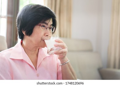 Happy Elderly People Health Care At Home Concept. Senior Asian Woman Drinking A Milk Glass Wear Pink Shirt In Living Room.