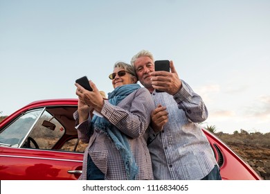 Happy elderly senior couple use mobile phones outdoor near a red beautiful retro car. leisure activity and travel together forever concept. modern use of technology from old adult that enjoyed life