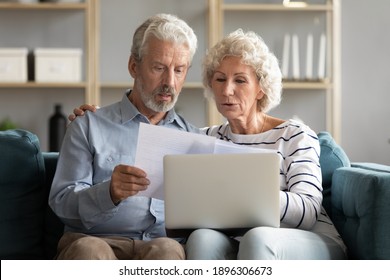 Happy elderly senior family couple reading paper correspondence or mails, sitting on sofa with computer on laps. Smiling mature spouses managing financial affairs indoors using laptop in living room.
