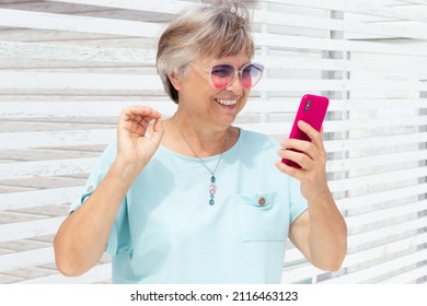 Happy Elderly Woman In Colored Sunglasses And A Blue T-shirt Uses A Smartphone For Video Chat