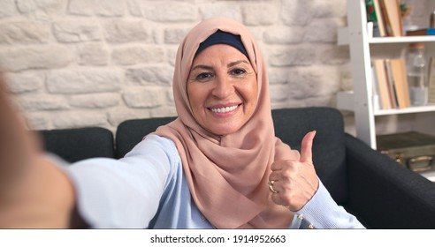 Happy Elderly Woman In Hijab Is Taking Selfie While Holding A Mobile Phone. Old Lady In A Headscarf Makes A Thumbs Up Sign. 