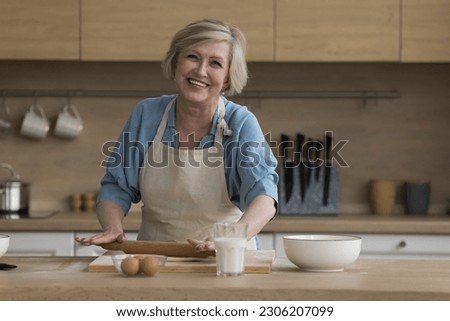 Happy elderly woman preparing pastries in kitchen, attractive mature female wear apron hold rolling-pin flattening prepared dough makes home-made cake. Family recipe, natural ingredient for cookery
