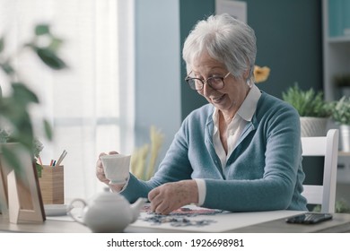 Happy elderly woman relaxing at home alone, she is solving a puzzle and drinking tea - Powered by Shutterstock