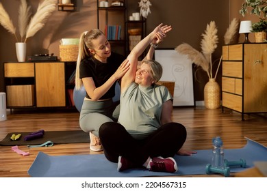 Happy elderly woman sitting on yoga mat looking at young adorable nurse helper doing stretching together in lotos pose in modern light room. Young girl helping senior lady reabiltaton process. - Powered by Shutterstock