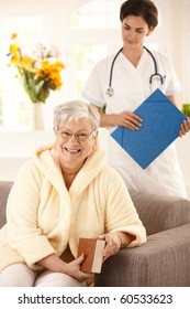 Happy Elderly Woman Sitting On Sofa At Home, Nurse Watching From Behind.?