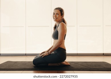 Happy elderly woman smiling at the camera while kneeling in thunderbolt pose. Mature woman doing a meditation exercise at home. Cheerful senior woman practicing hatha yoga on an exercise mat. - Powered by Shutterstock
