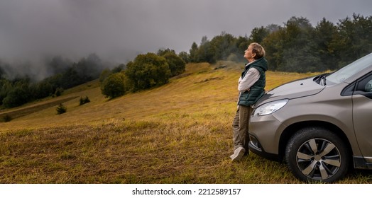 Happy Elderly Woman Traveler Standing Near The Car While Traveling In The Mountains. Active Retirees Travel Lifestyle