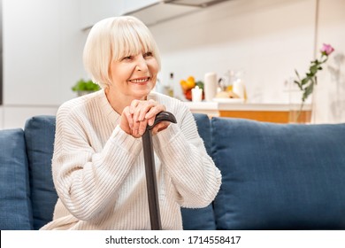 Happy elderly woman with walking cane in hands spending weekend at home, sitting on couch in living room, looking aside, making beaming smile - Shutterstock ID 1714558417