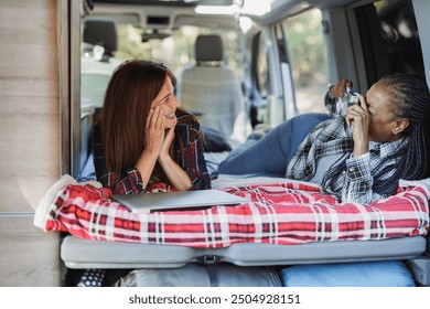 Happy elderly women having fun inside mini van camper during autumn vacation - African woman taking a photo form her caucasian female friend - Tavel concept - Focus on woman hand - Powered by Shutterstock