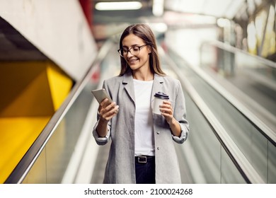 A happy, elegant businesswoman descending the escalator and holding takeaway coffee while reading important messages. A woman on the escalator with a phone - Powered by Shutterstock