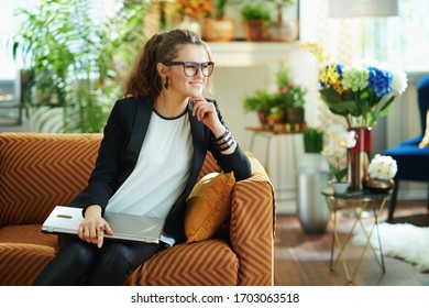 Happy Elegant Female In White Blouse And Black Jacket With Closed Laptop Sitting On Couch In The Modern House In Sunny Day.