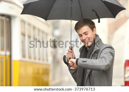 Happy elegant man consulting information in a smartwatch waiting for public transportation in the street of an old town under an umbrella in a rainy day