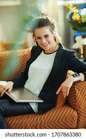 Happy Elegant Middle Age Woman In White Blouse And Black Jacket With Closed Laptop Sitting On Couch In The Modern House In Sunny Day.