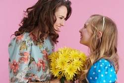 Happy Elegant Mother And Daughter With Long Wavy Hair Presenting Yellow Chrysanthemums Flowers Against Pink Background.
