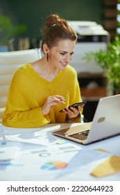 Happy Elegant Small Business Owner Woman In Yellow Sweater With Laptop Using Smartphone In The Modern Green Office.