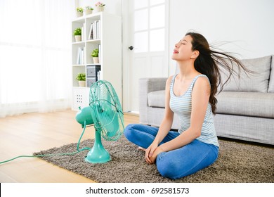 Happy Elegant Woman Blowing Electric Fan In Summer Day And Sitting On Living Room Floor Enjoying Cool Wind To Refreshing.