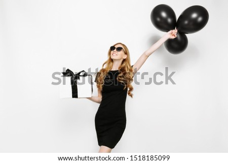 Happy elegant young girl in black dress and sunglasses, happily holding a gift with black Friday ribbon and black balloons on white background. Shopping, discounts, Black Friday