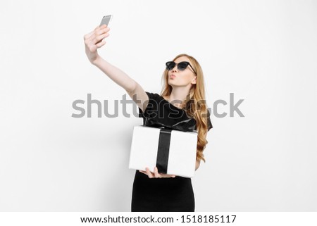 Happy elegant young girl in a black dress and sunglasses, holding a gift with a black ribbon black Friday, takes a selfie on a smartphone, on a white background. Shopping, discounts, Black Friday