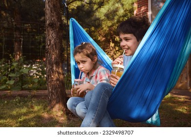 Happy elementary age kids have fun together, smiling, relaxing in blue hammock at summer camp, drinking healthy smoothie from straw. Children. Childhood. Family leisure and relationships. Friendship. - Powered by Shutterstock