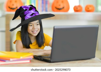 Happy Elementary Child In Halloween Costume.Asian Girl Online Learning.Kid With Purple And Black Witch Hat Using Laptop For Remote Lesson.Homeschooling.Pumpkin And Books In Background.