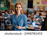 Happy elementary school teacher using touchpad in the classroom and looking at camera. Her students are learning in the background.