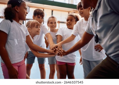 Happy elementary students joining hands in unity with their sports teacher at school gym.