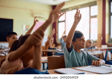 Happy elementary students raising their arms to answer teacher's question in the classroom. Focus is on boy in blue shirt. - Powered by Shutterstock