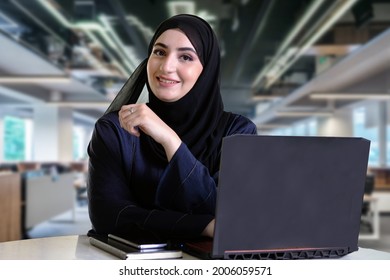 Happy Emirati woman on Abaya lookikng at the front camera with computer laptop, notebook and blurred office - Shutterstock ID 2006059571