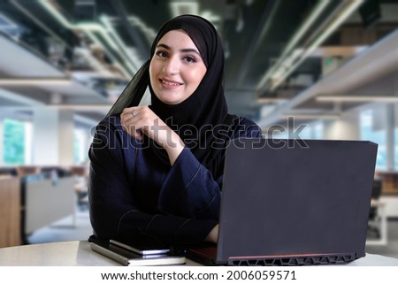Happy Emirati woman on Abaya lookikng at the front camera with computer laptop, notebook and blurred office